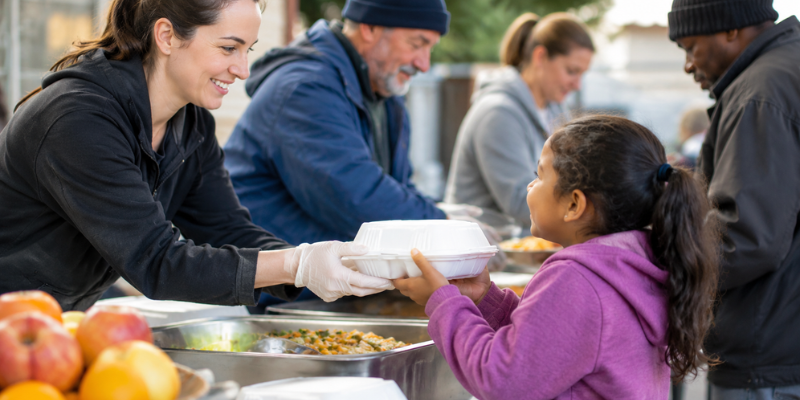 A volunteer offering a warm packaged meal container to a smiling child as part of meal sponsorship for dignity program.