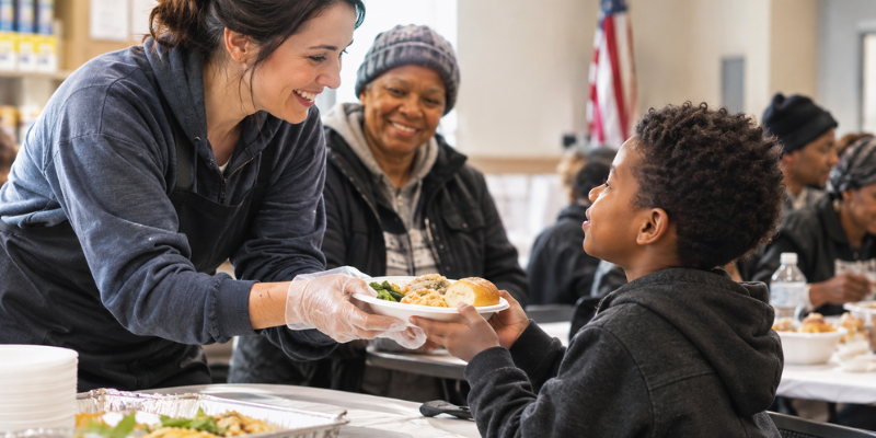 A cheerful volunteer serving a warm plate of food to a young boy, representing food as comfort and care in a community center.